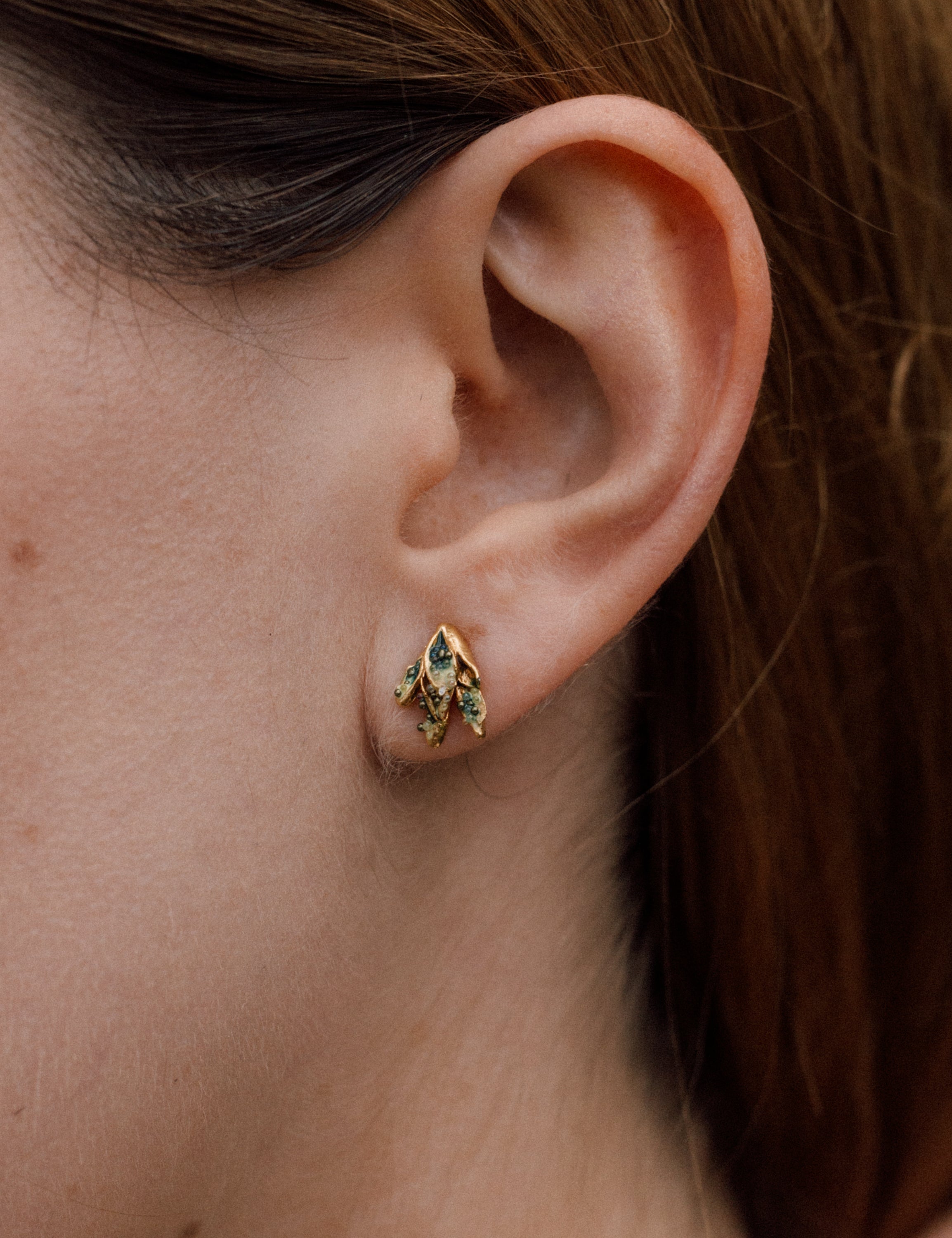 Close-up side view of a woman’s ear wearing a single Topos Nebula Mini stud earring in gold-plated brass, hand-painted with green  paint and glass granulation, her hair tucked behind her ear against a neutral backdrop.