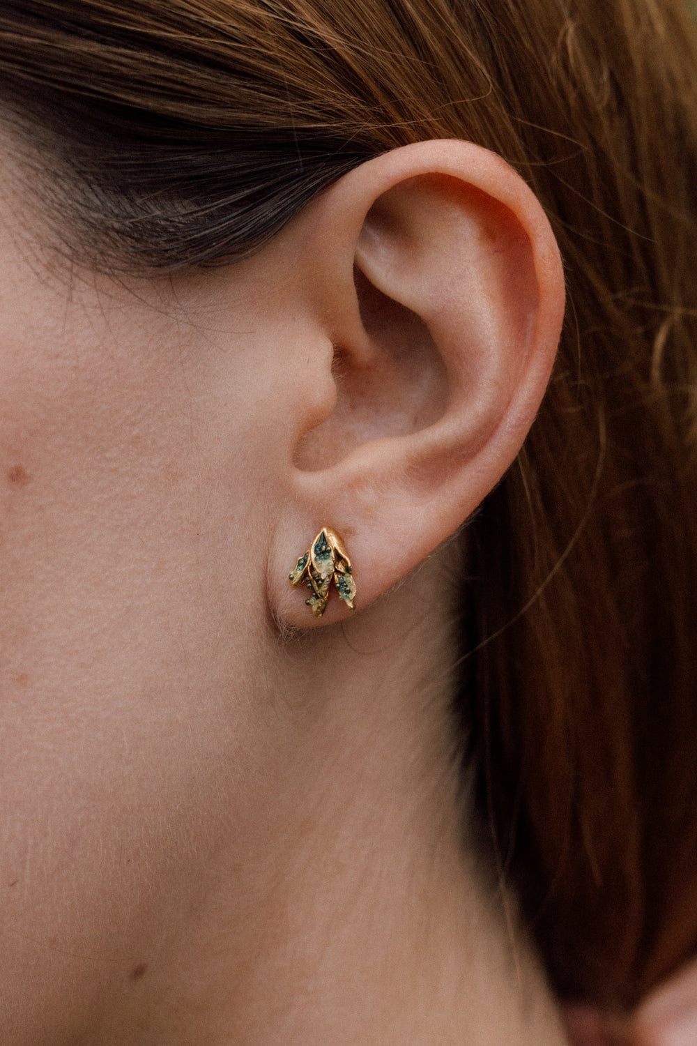 Close-up side view of a woman’s ear wearing a single Topos Nebula Mini stud earring in gold-plated brass, hand-painted with green  paint and glass granulation, her hair tucked behind her ear against a neutral backdrop.