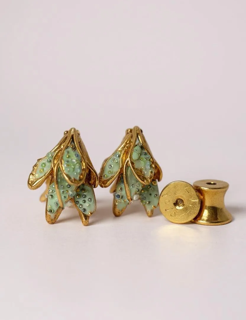 Flat lay of two Topos Nebula Mini leaf-shaped stud earrings hand-painted in green with glass granulation, accompanied by a separate round gold-plated earring back on a white surface