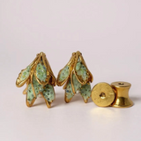 Flat lay of two Topos Nebula Mini leaf-shaped stud earrings hand-painted in green with glass granulation, accompanied by a separate round gold-plated earring back on a white surface