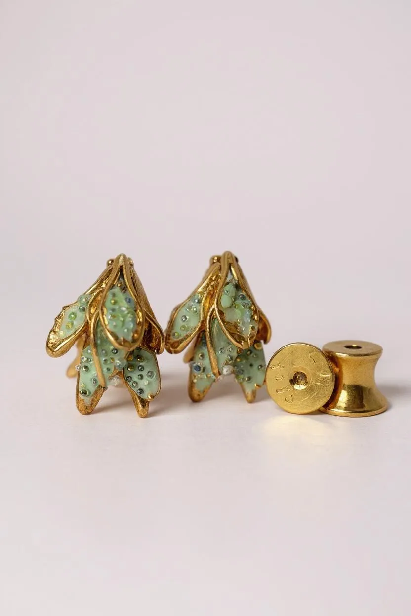 Flat lay of two Topos Nebula Mini leaf-shaped stud earrings hand-painted in green with glass granulation, accompanied by a separate round gold-plated earring back on a white surface