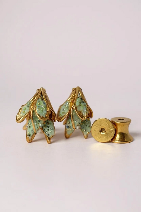Flat lay of two Topos Nebula Mini leaf-shaped stud earrings hand-painted in green with glass granulation, accompanied by a separate round gold-plated earring back on a white surface