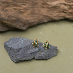 Two Topos Nebula Mini leaf-shaped studs with gold-plated bronze settings, hand-painted in green and finished with glass granulation, resting on a grey stone against an olive-green background.