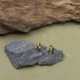 Two Topos Nebula Mini leaf-shaped studs with gold-plated bronze settings, hand-painted in green and finished with glass granulation, resting on a grey stone against an olive-green background.