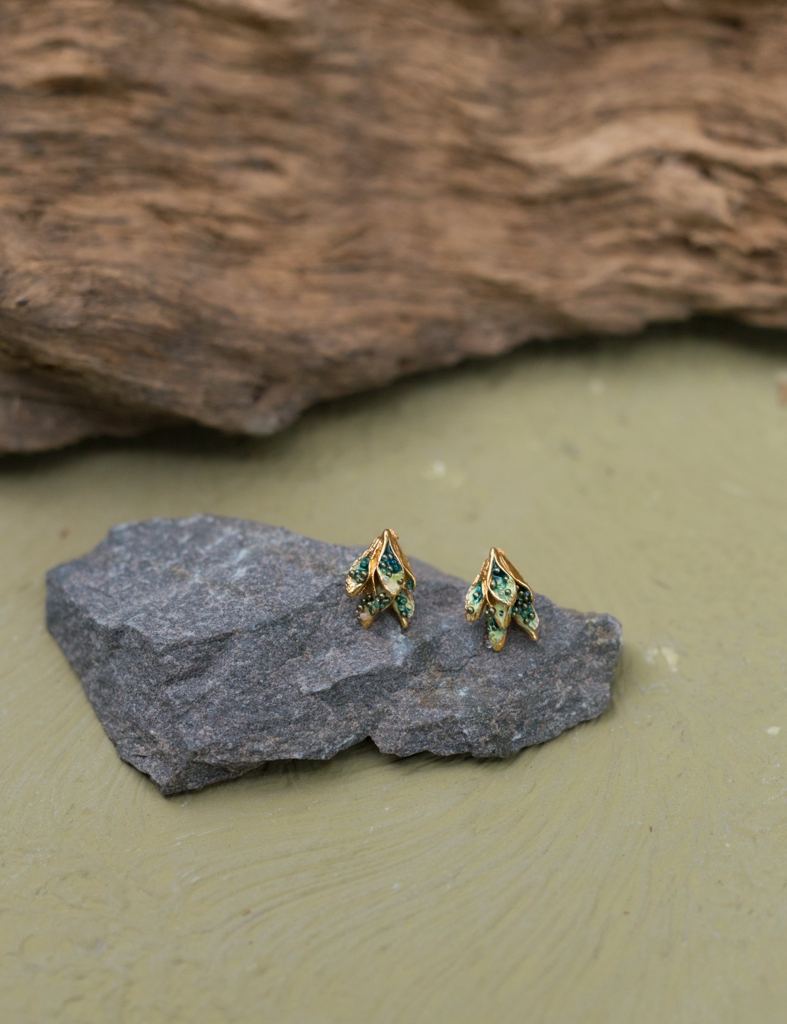 Two Topos Nebula Mini leaf-shaped studs with gold-plated bronze settings, hand-painted in green and finished with glass granulation, resting on a grey stone against an olive-green background.