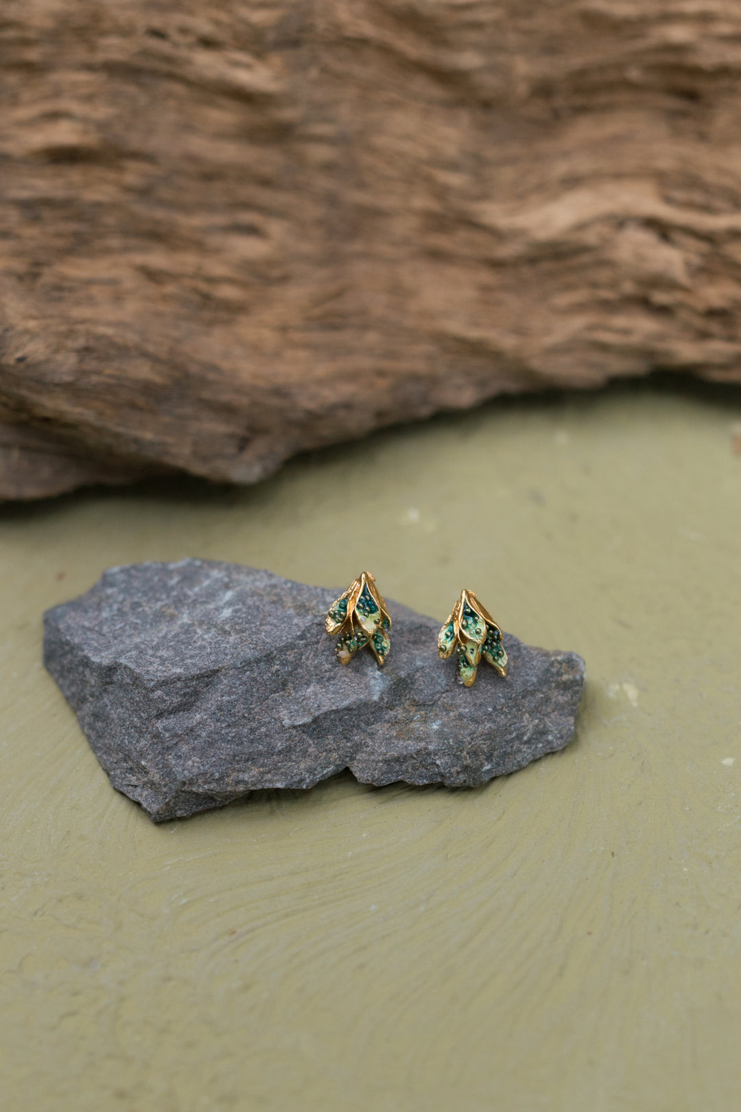 Two Topos Nebula Mini leaf-shaped studs with gold-plated bronze settings, hand-painted in green and finished with glass granulation, resting on a grey stone against an olive-green background.