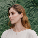 Portrait of a woman wearing a white blouse and a Topos Nebula Mini stud earring in gold-plated brass, hand-painted in green  with glass granulation, against a backdrop of large green leaves.