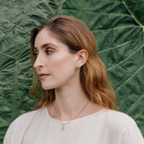 Portrait of a woman wearing a white blouse and a Topos Nebula Mini stud earring in gold-plated brass, hand-painted in green  with glass granulation, against a backdrop of large green leaves.