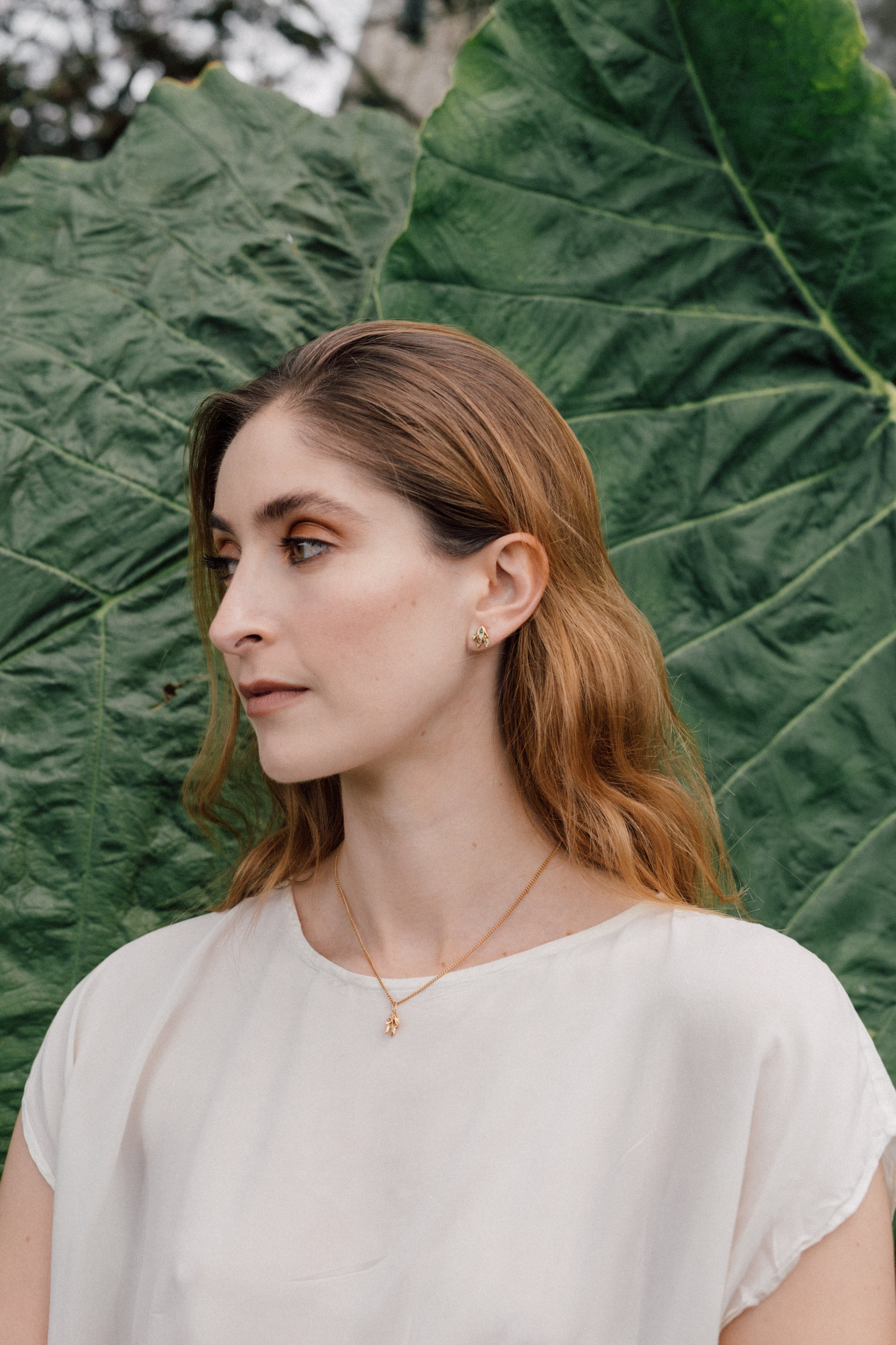 Portrait of a woman wearing a white blouse and a Topos Nebula Mini stud earring in gold-plated brass, hand-painted in green  with glass granulation, against a backdrop of large green leaves.