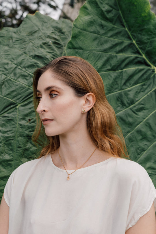 Portrait of a woman wearing a white blouse and a Topos Nebula Mini stud earring in gold-plated brass, hand-painted in green  with glass granulation, against a backdrop of large green leaves.