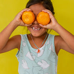 “Young girl in a mint sleeveless top holds two oranges over her eyes, smiling against a bright yellow background.