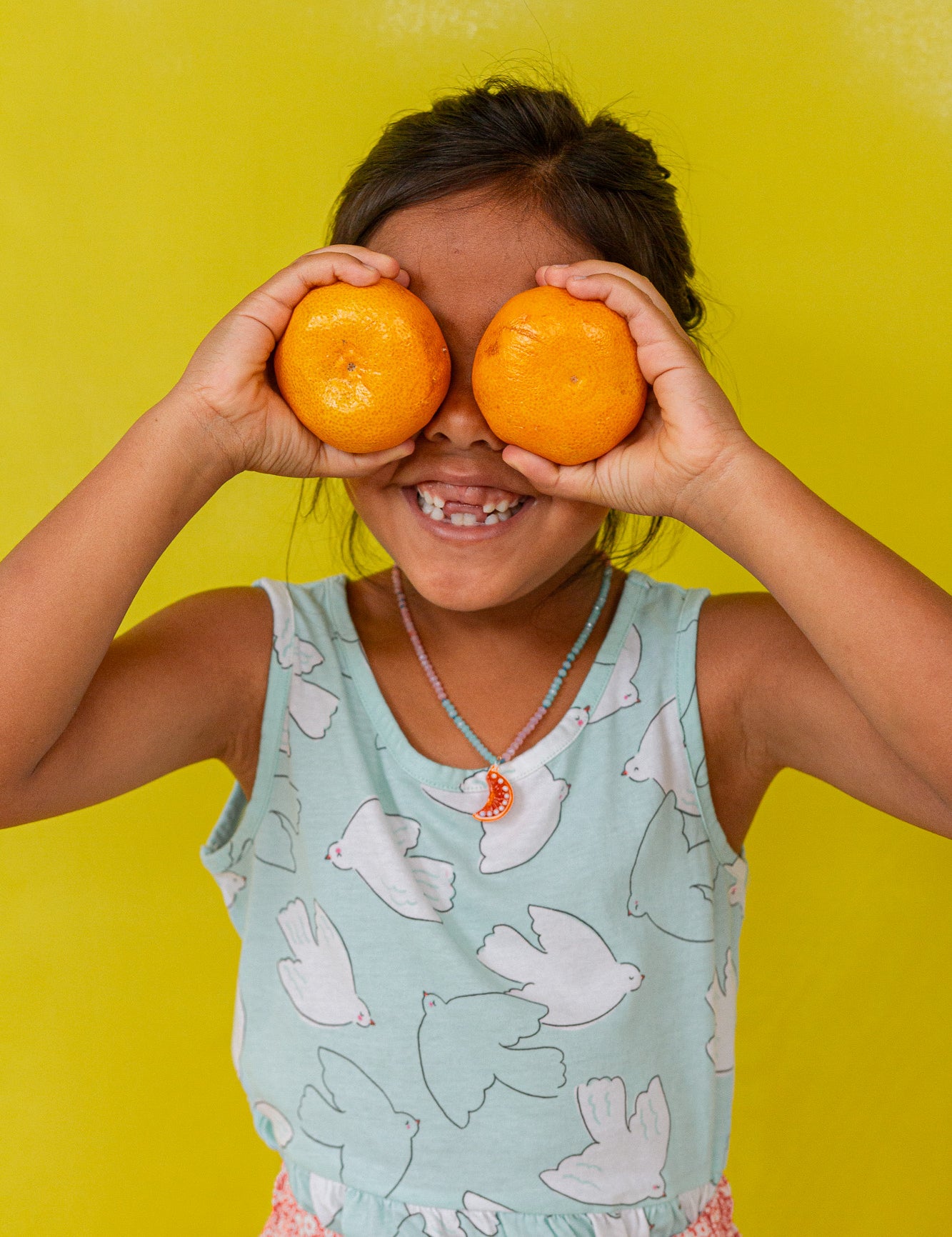 “Young girl in a mint sleeveless top holds two oranges over her eyes, smiling against a bright yellow background.