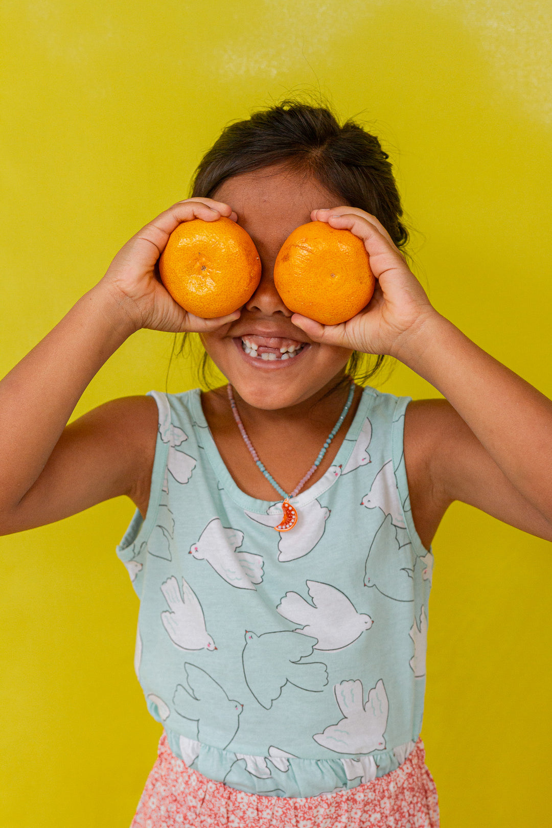 “Young girl in a mint sleeveless top holds two oranges over her eyes, smiling against a bright yellow background.