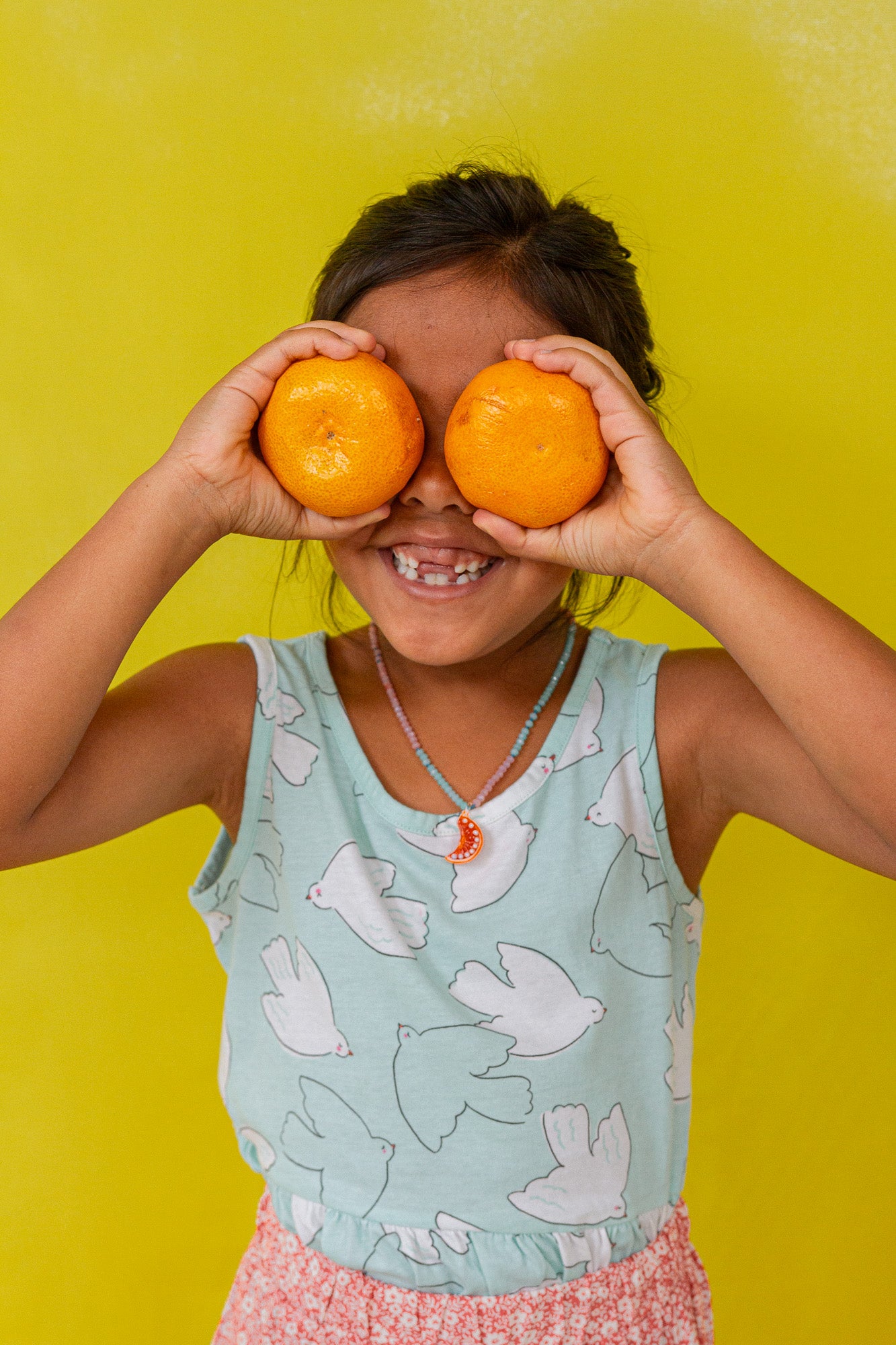 “Young girl in a mint sleeveless top holds two oranges over her eyes, smiling against a bright yellow background.