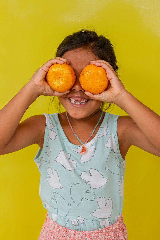 “Young girl in a mint sleeveless top holds two oranges over her eyes, smiling against a bright yellow background.