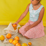 Child kneeling on the floor in a mint top and pastel skirt plays with oranges and wears the Orange Necklace.