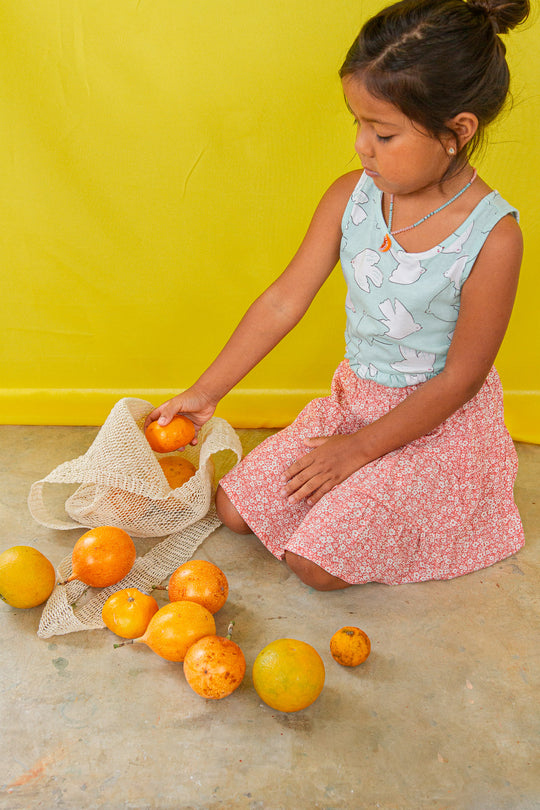 Child kneeling on the floor in a mint top and pastel skirt plays with oranges and wears the Orange Necklace.