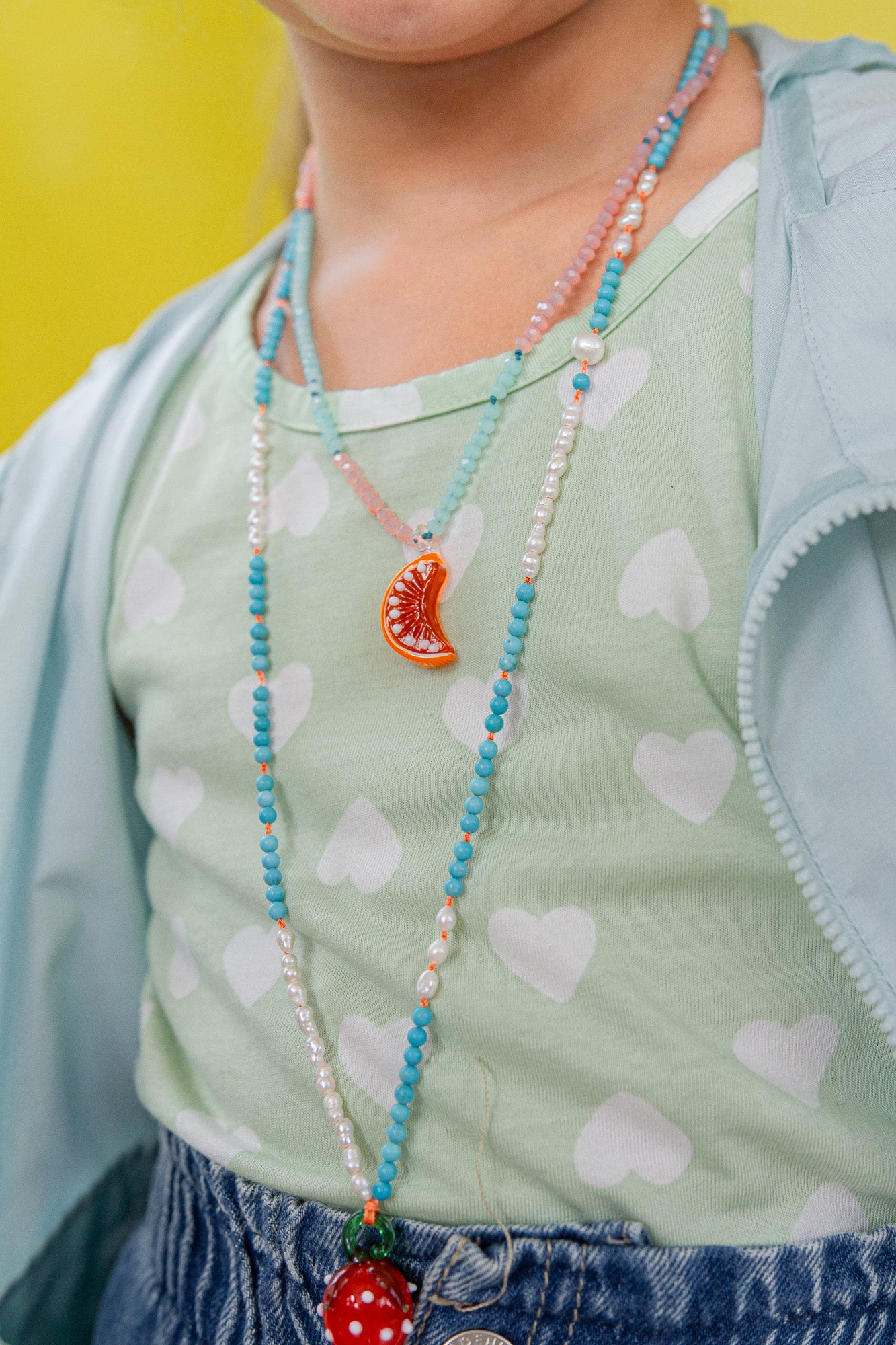 Close‑up of a child in a mint sleeveless top wearing the Orange Necklace, with the pendant resting near her chest against a yellow background