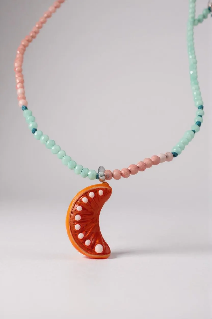 Close-up of the Orange Necklace pendant and beads, highlighting the glossy bright orange crescent moon charm decorated with tiny metal studs amid mint-green and pink beads