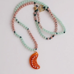 Orange Necklace coiled in a loose spiral on a grey background, showing multiple layers of alternating mint-green and pink beads with the bright orange crescent moon pendant peeking out.