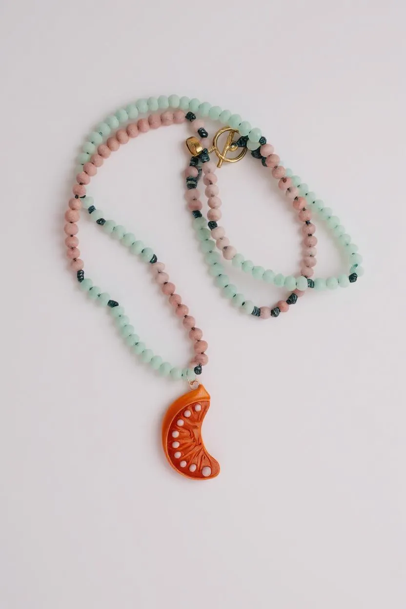 Orange Necklace coiled in a loose spiral on a grey background, showing multiple layers of alternating mint-green and pink beads with the bright orange crescent moon pendant peeking out.
