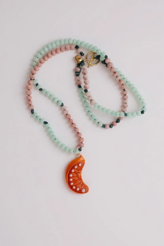 Orange Necklace coiled in a loose spiral on a grey background, showing multiple layers of alternating mint-green and pink beads with the bright orange crescent moon pendant peeking out.