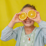 “Child wearing a light denim jacket and mint top holds two oranges over her eyes against a yellow backdrop.