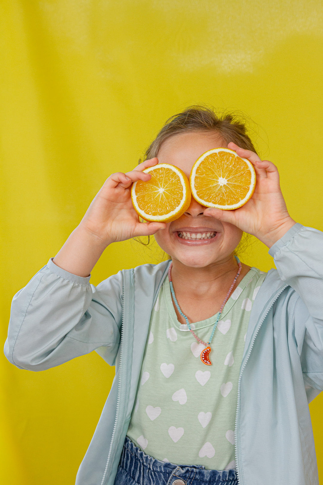 “Child wearing a light denim jacket and mint top holds two oranges over her eyes against a yellow backdrop.