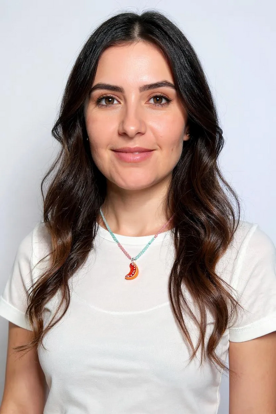 Portrait of a woman with long dark hair wearing a white shirt and the Orange Necklace, centered on her neckline against a white background.