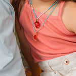 Close‑up of a child’s torso wearing a coral top and white shorts, featuring the Orange Necklace pendant at chest level.