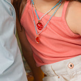 Close‑up of a child’s torso wearing a coral top and white shorts, featuring the Orange Necklace pendant at chest level.