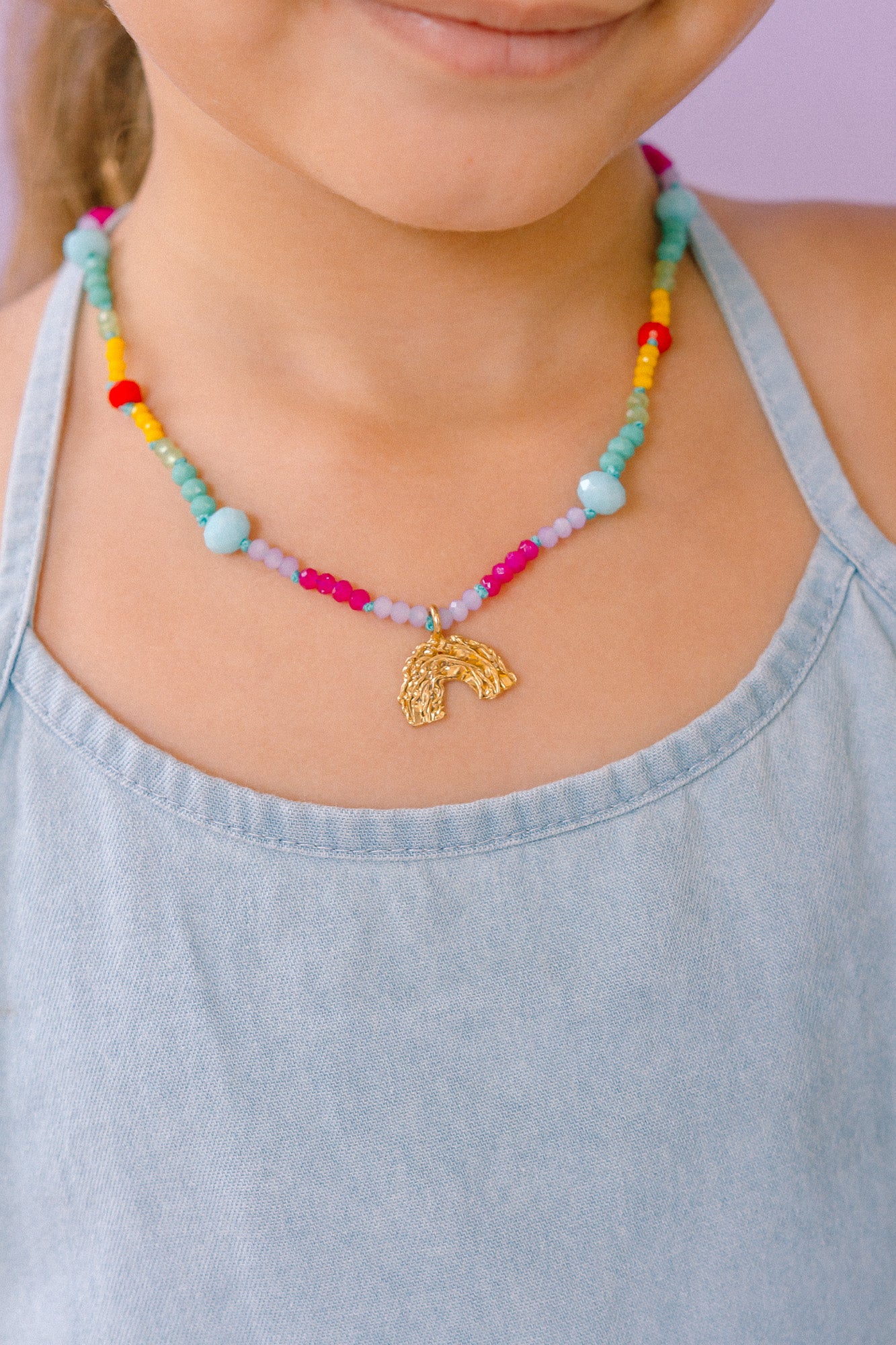 Multicolored beaded necklace with a gold rainbow-shaped pendant, worn by a girl wearing a light blue top.