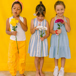 Three girls wearing multicolored beaded necklaces, holding flowers against a yellow background.