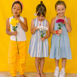 Three girls wearing multicolored beaded necklaces, holding flowers against a yellow background.
