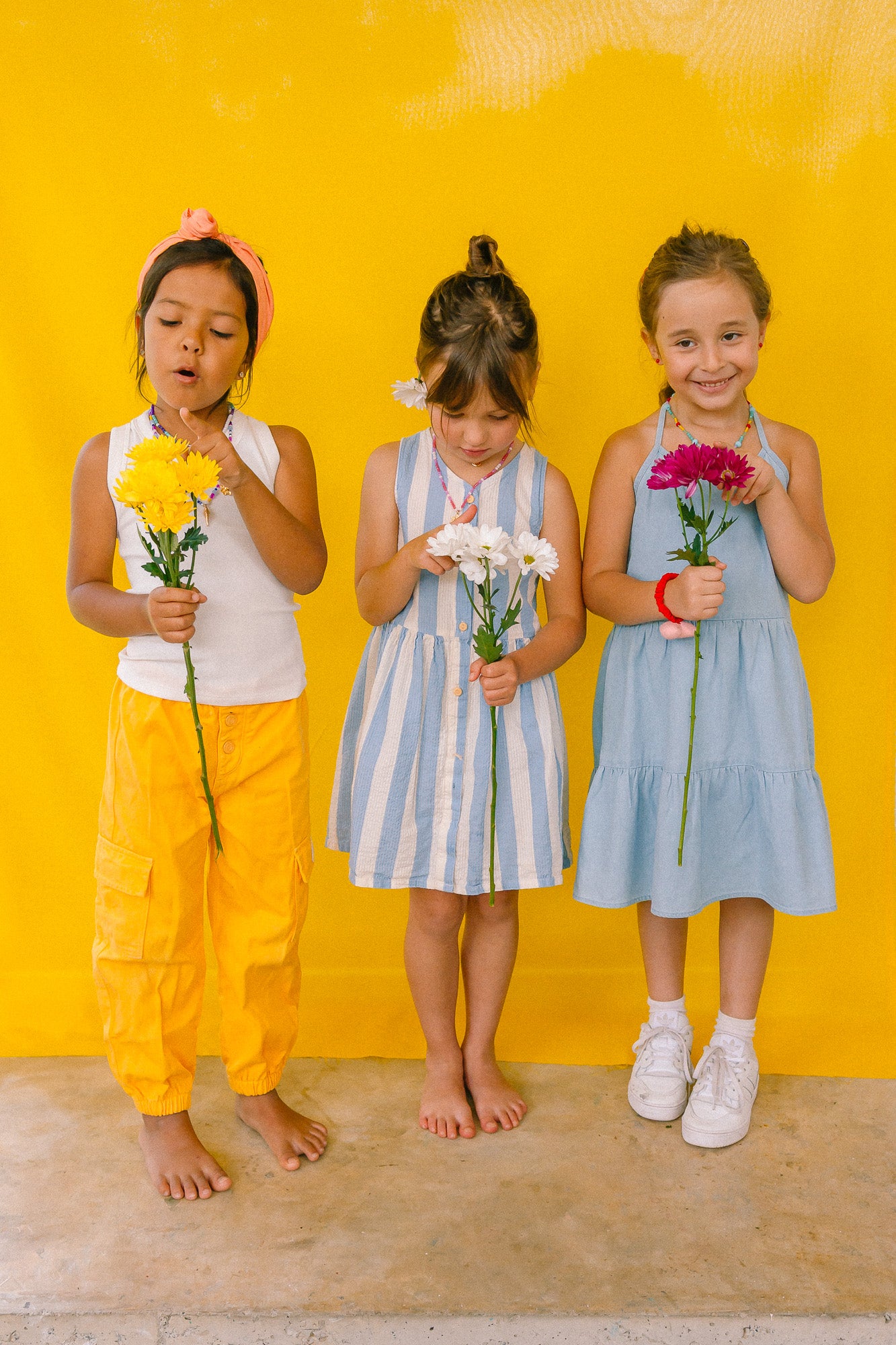 Three girls wearing multicolored beaded necklaces, holding flowers against a yellow background.