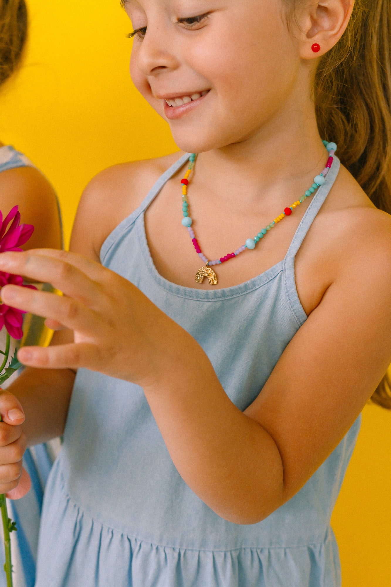 Girl in half profile wearing a colorful necklace and holding a pink flower on a yellow background.