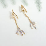 Gold-plated brass Root earrings with lavender tips and glass granulation, photographed against a blurred botanical background.