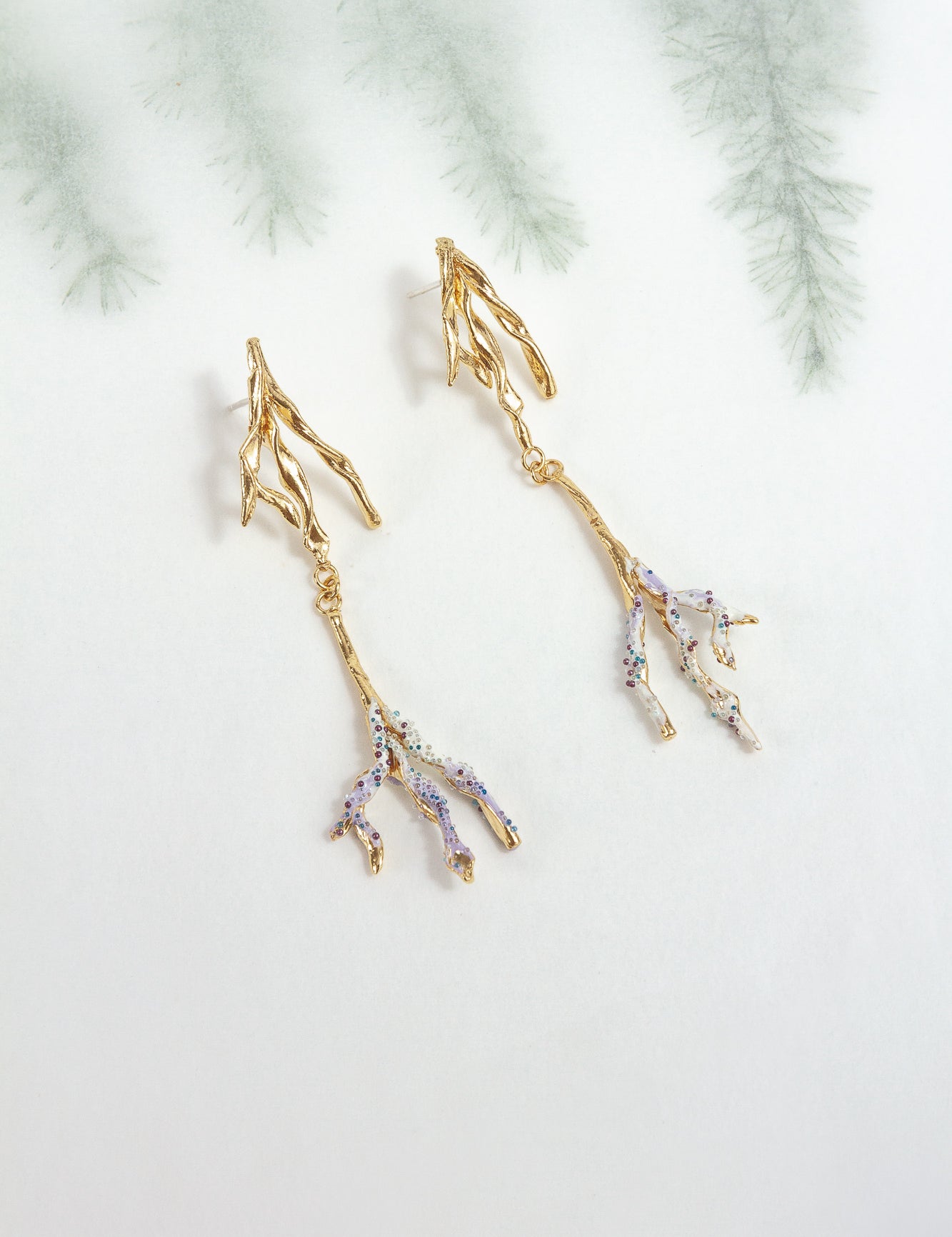 Gold-plated brass Root earrings with lavender tips and glass granulation, photographed against a blurred botanical background.