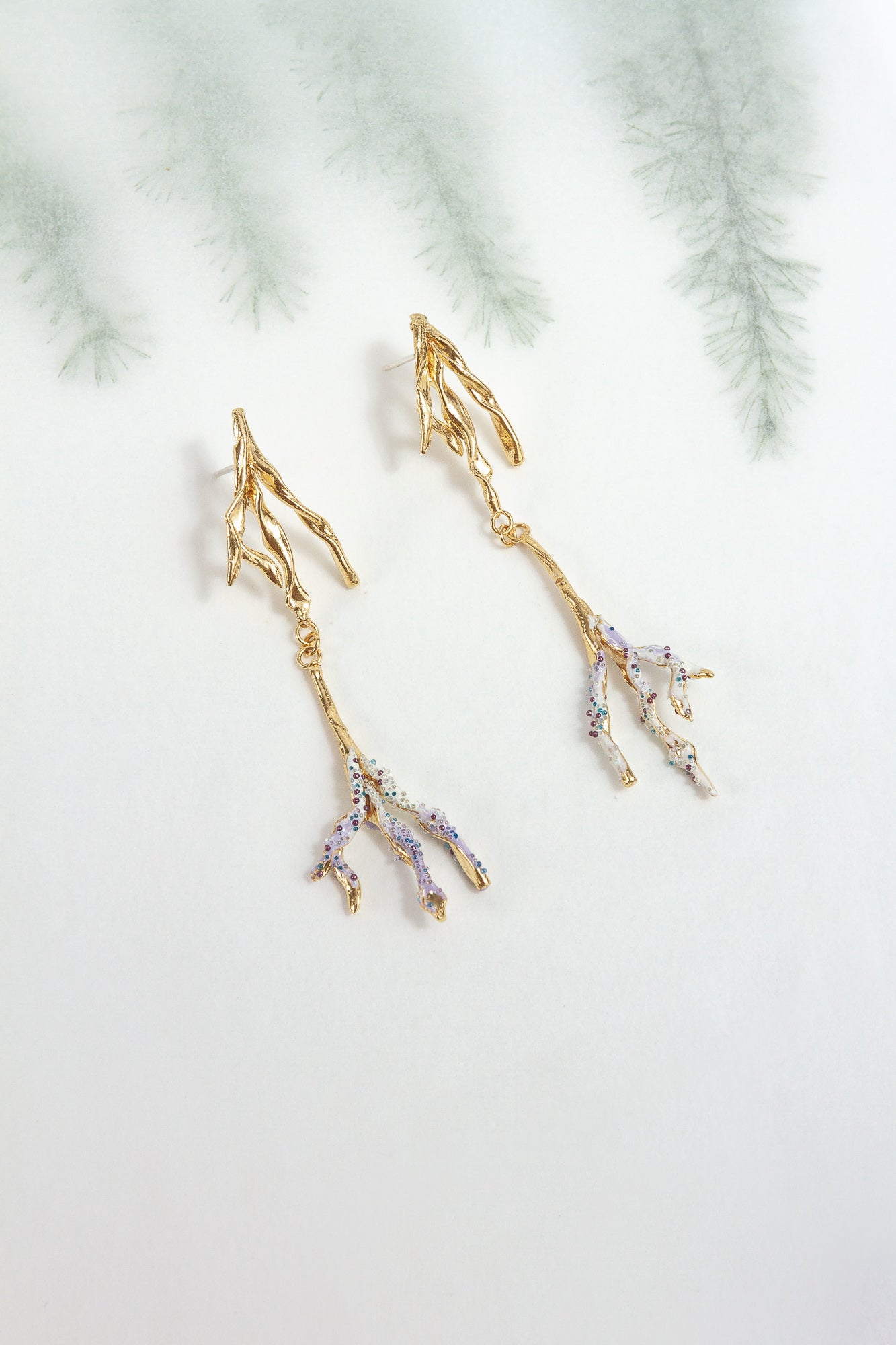 Gold-plated brass Root earrings with lavender tips and glass granulation, photographed against a blurred botanical background.