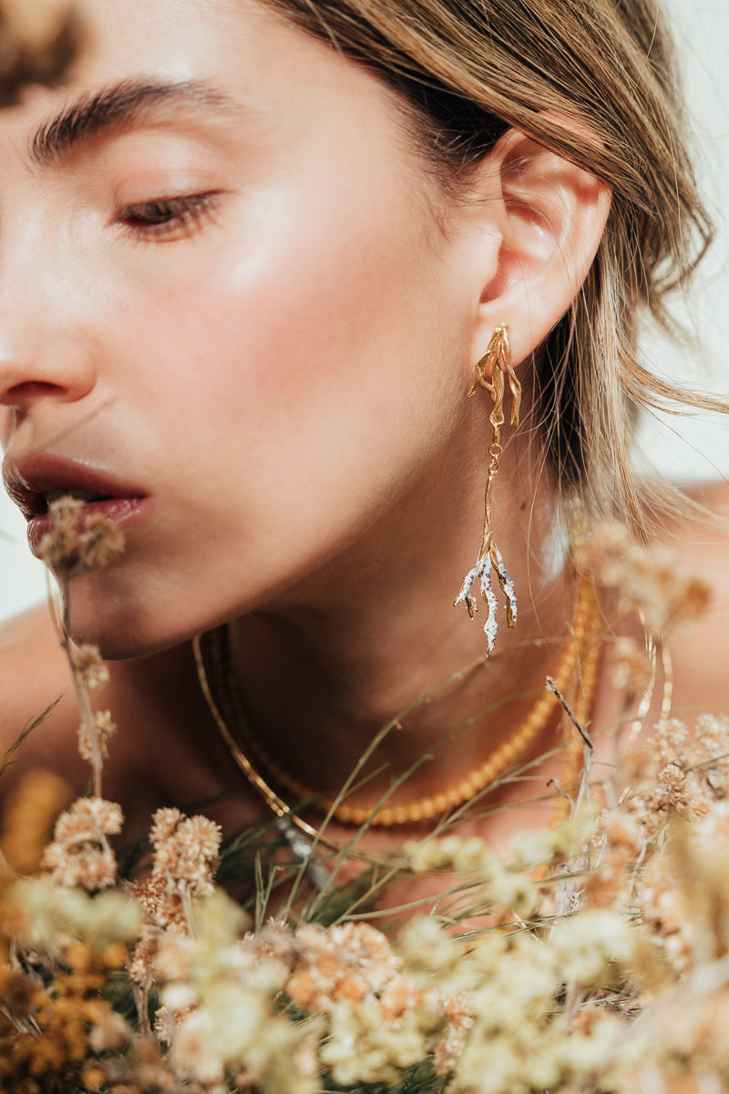 Close-up of a model wearing the Root Earrings, in an organic setting surrounded by dried plants and earth tones.