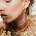 Close-up of a model wearing the Root Earrings, in an organic setting surrounded by dried plants and earth tones.
