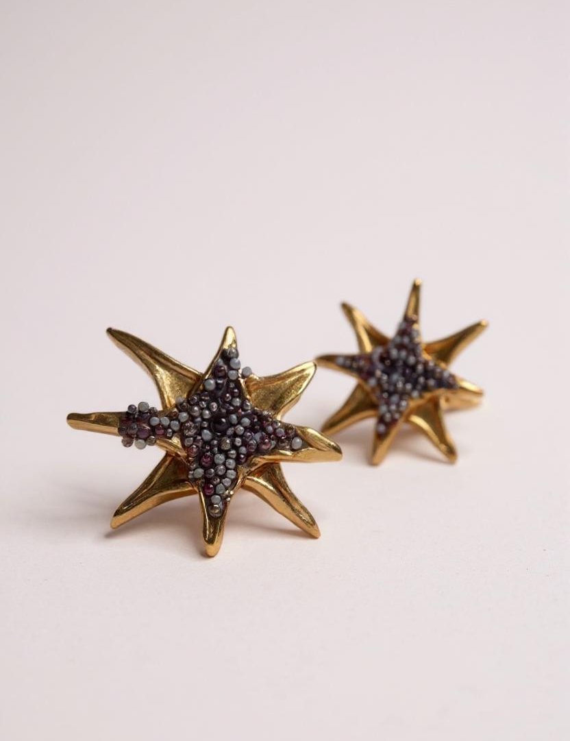 Pair of gold-tone star-shaped stud earrings encrusted with small rust crystals, displayed on a pale pink background.