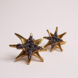 Pair of gold-tone star-shaped stud earrings encrusted with small rust crystals, displayed on a pale pink background.