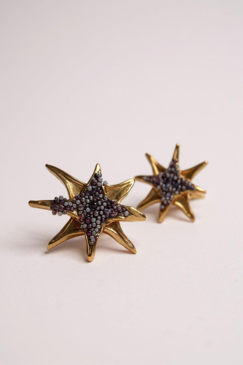 Pair of gold-tone star-shaped stud earrings encrusted with small rust crystals, displayed on a pale pink background.