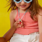 Girl wearing white‑rimmed sunglasses holds the Strawberry Necklace pendant near her face against a yellow backdrop.
