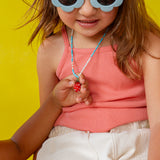 Girl wearing white‑rimmed sunglasses holds the Strawberry Necklace pendant near her face against a yellow backdrop.
