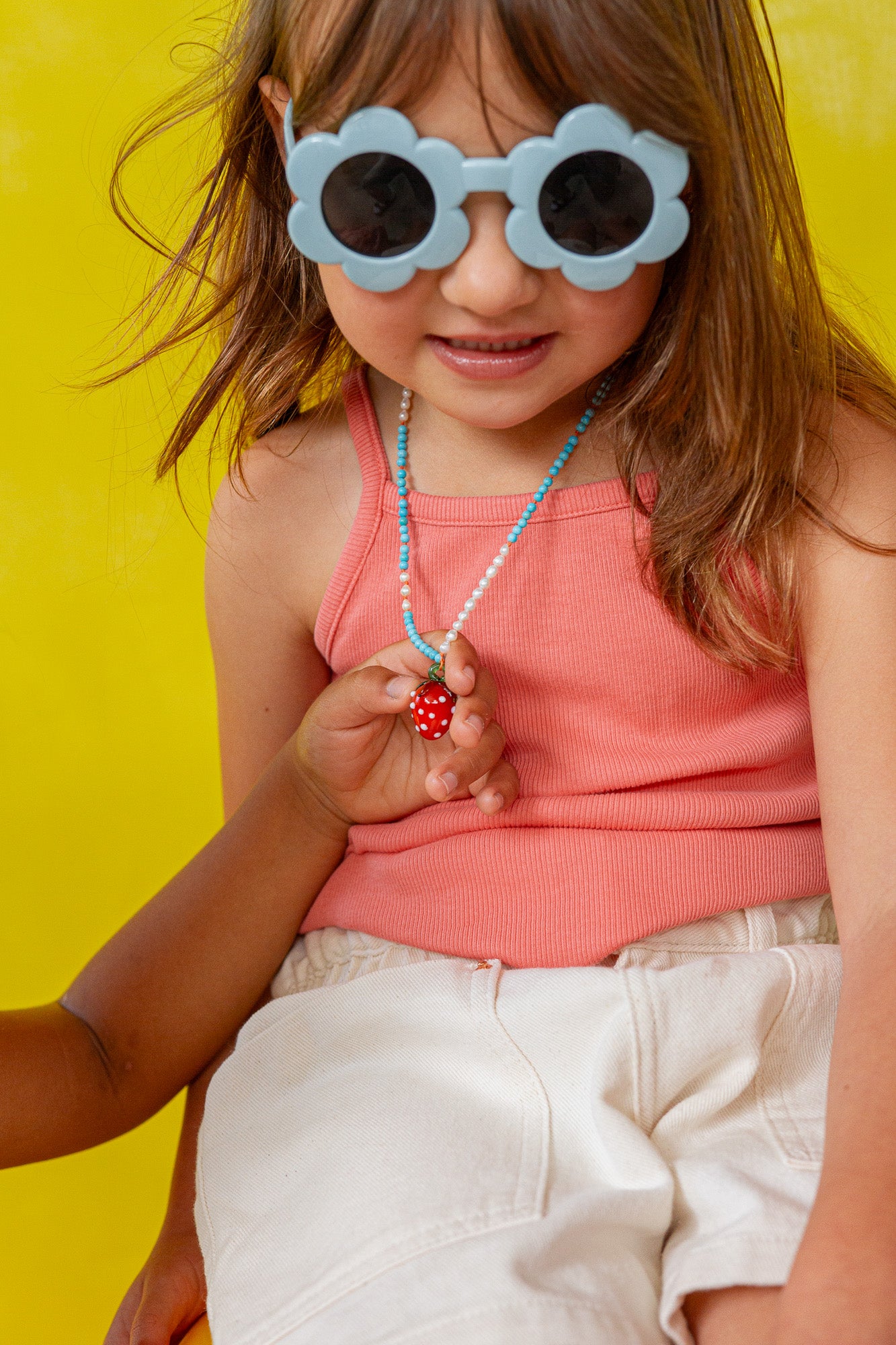 Girl wearing white‑rimmed sunglasses holds the Strawberry Necklace pendant near her face against a yellow backdrop.