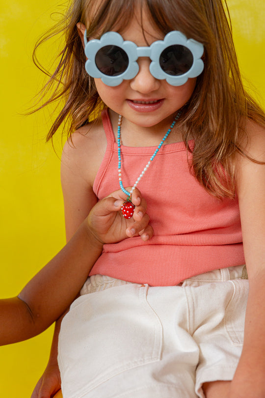 Girl wearing white‑rimmed sunglasses holds the Strawberry Necklace pendant near her face against a yellow backdrop.