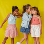 Full‑length portrait of three girls in colorful summer outfits and the Strawberry Necklace, posing playfully against a yellow background.
