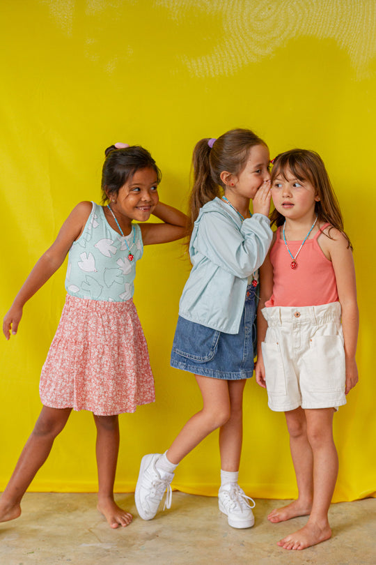 Full‑length portrait of three girls in colorful summer outfits and the Strawberry Necklace, posing playfully against a yellow background.